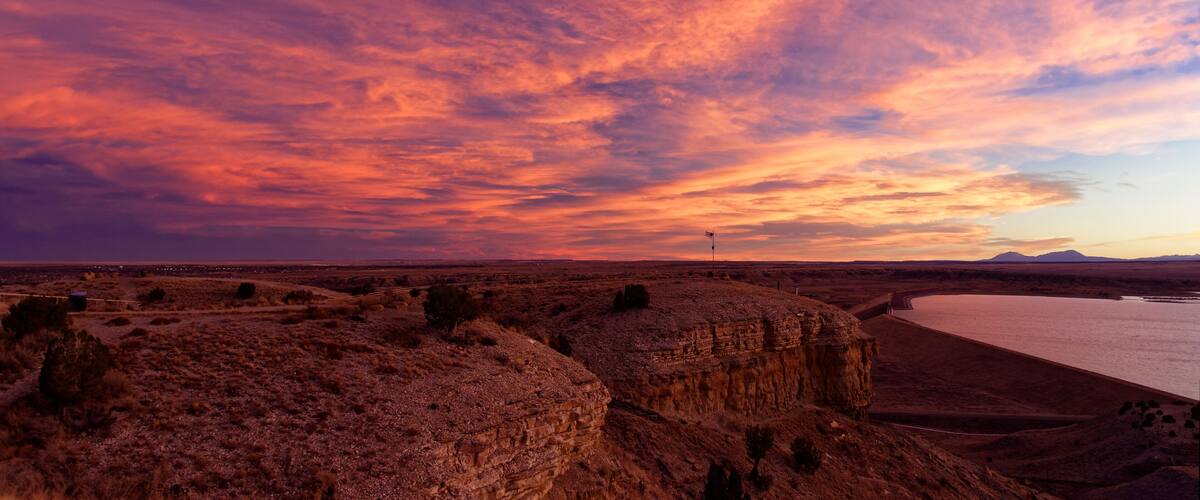 A desert landscape with a body of water, rocky formations, and a sunset sky.