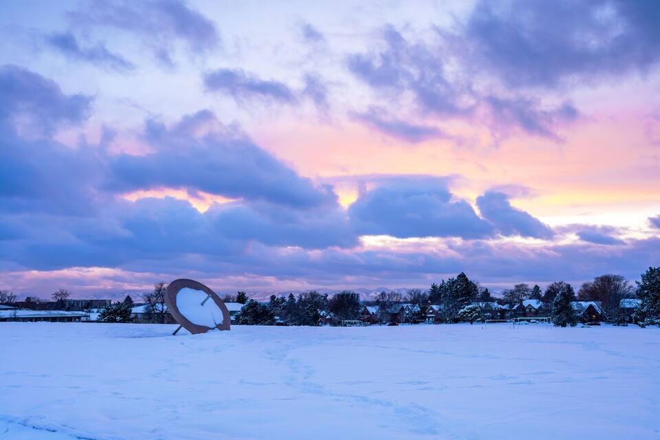 Paisaje nevado con una antena parabólica en primer plano y casas al fondo bajo un cielo colorido.