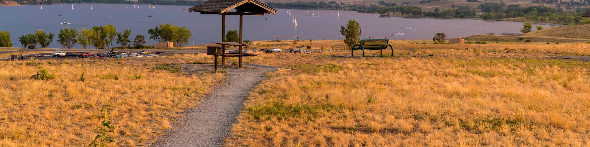 Un campo cubierto de hierba con un pabellón de madera, un banco y un sendero que conduce a un lago con veleros.