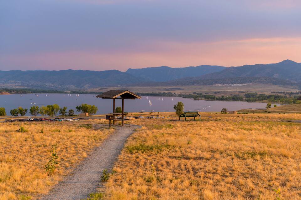 A grassy field with a wooden pavilion, a bench, and a path leading to a lake with sailboats.