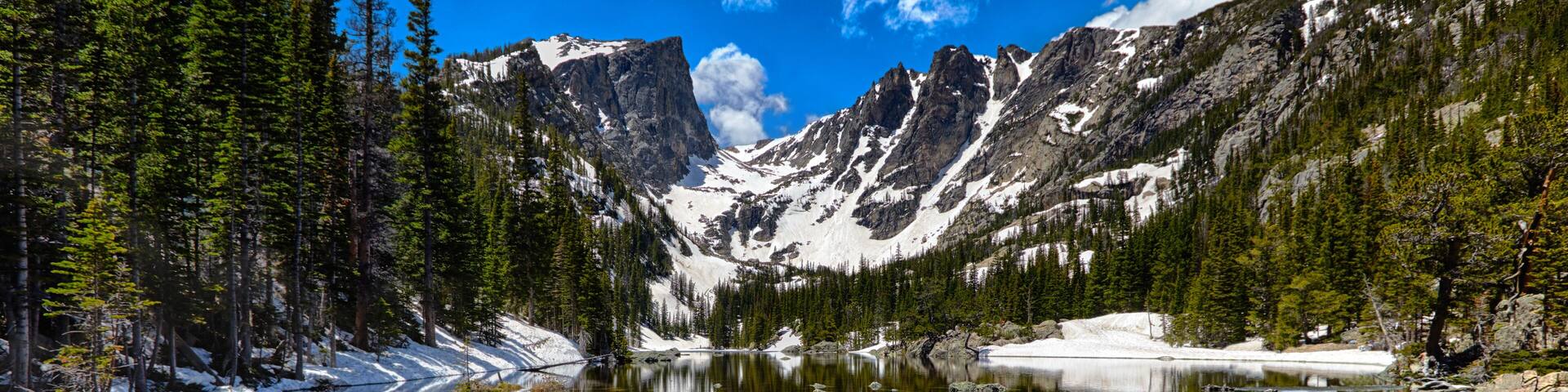 A clear mountain lake surrounded by snow-capped peaks and dense evergreen forests.