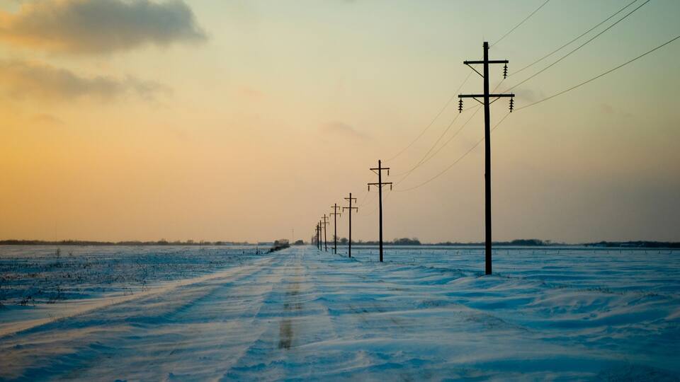 A snow-covered road with utility poles and power lines extending into the distance.