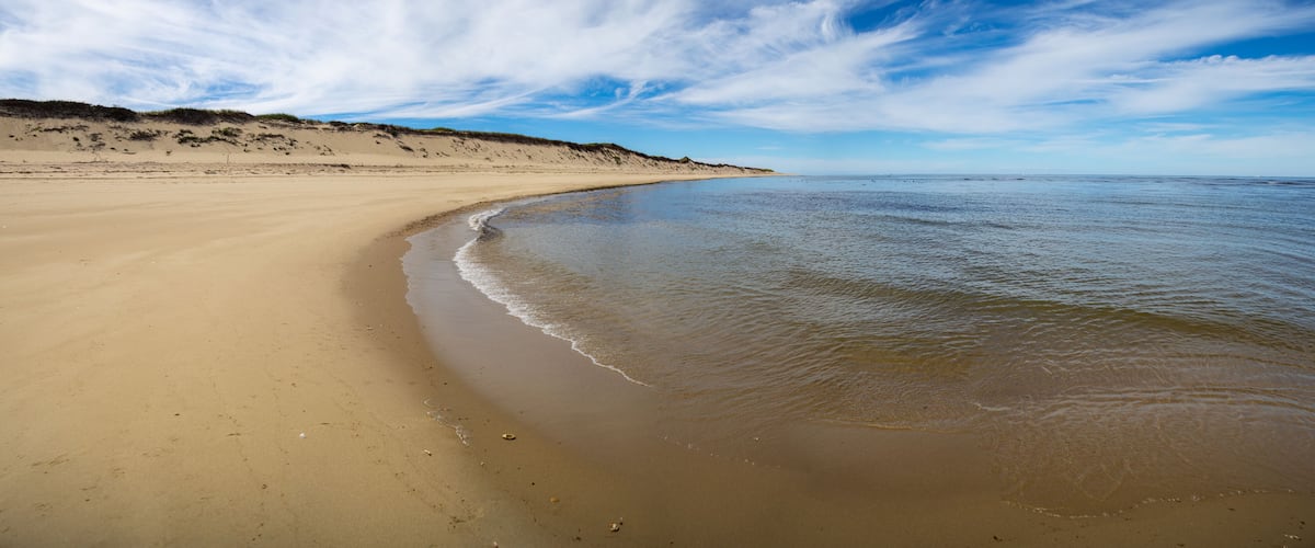 Een zandstrand met rustige golven, een heldere lucht en duinen in de verte.