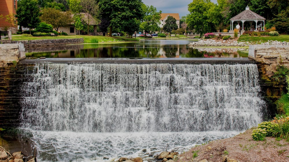 Ein Wasserfall stürzt in einen Teich, der von viel Grün und einem Pavillon umgeben ist.