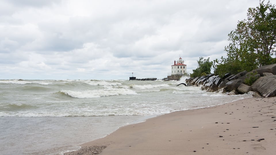 Lighthouse at Mentor Headlands Beach State Park in Ohio with waves crashing on shore and dark clouds overhead.