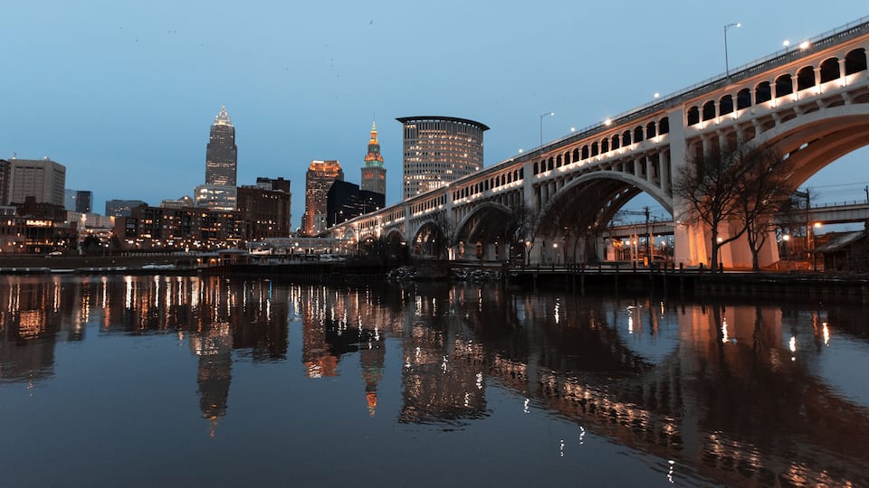 A city skyline with illuminated buildings, a bridge with arches, and a calm river reflecting the lights.