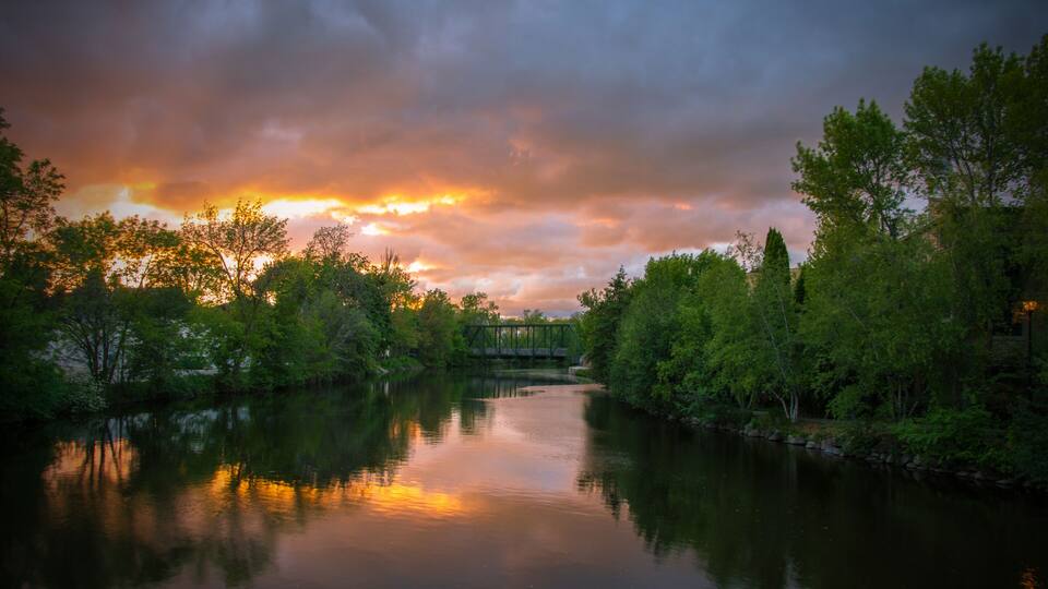 A river reflecting the sunset with trees on both sides and a bridge in the distance.