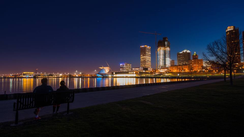 A nighttime cityscape with a river reflecting lights, a modern building with a unique roof, and a person sitting on a bench.