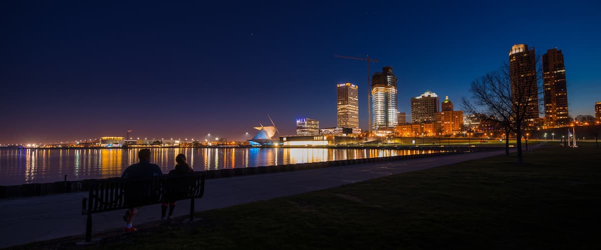 A nighttime cityscape with a river reflecting lights, a modern building with a unique roof, and a person sitting on a bench.