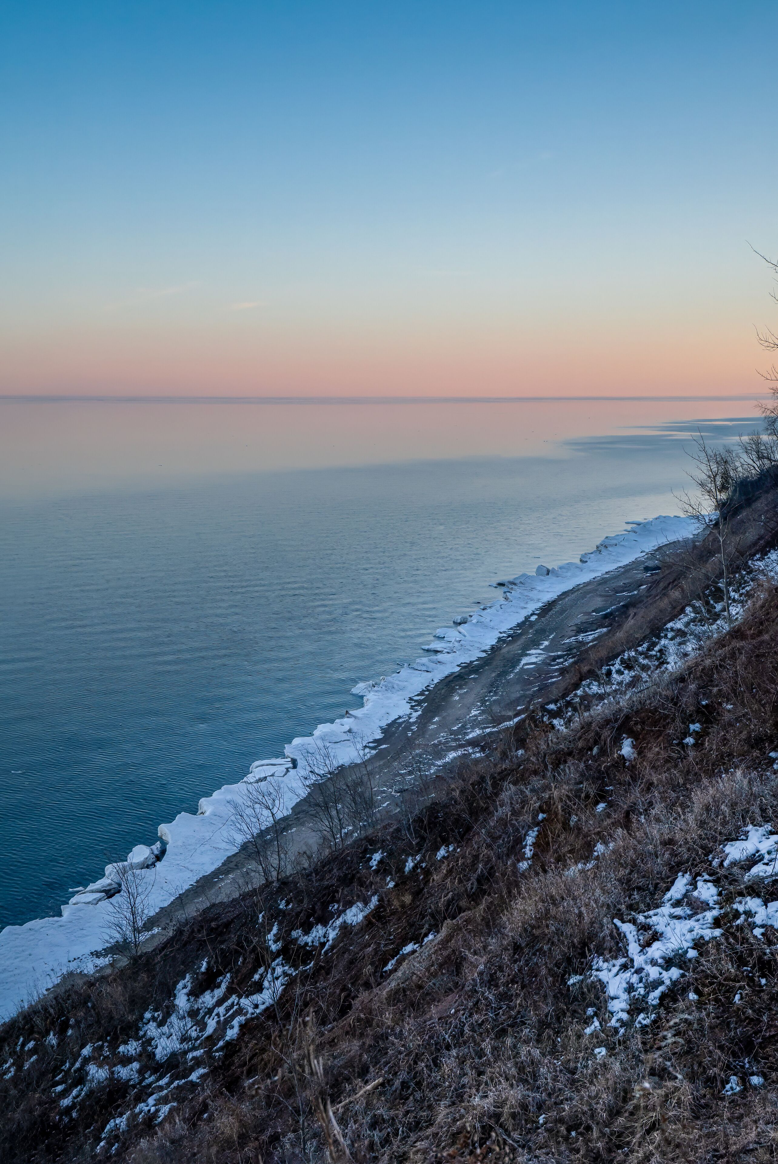 Scenic winter sunset over Lake Michigan at Lion's Den Gorge Nature Preserve in Wisconsin. High bluffs overlook an icy shoreline with a pink and blue twilight horizon. 
Spring is coming.