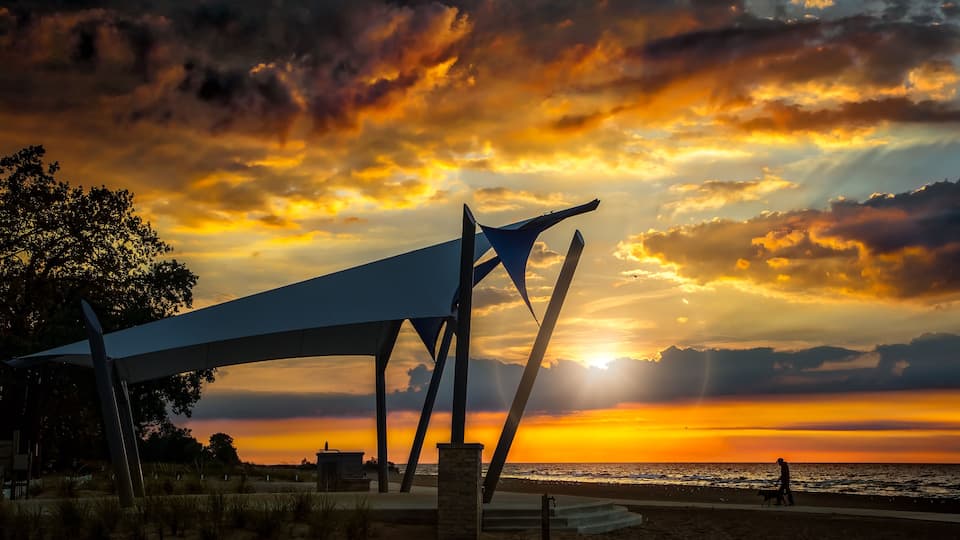 A sunset over a beach with a modern sculpture and a person walking a dog.