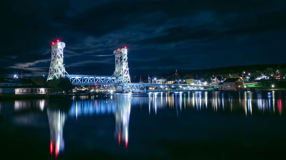 A nighttime view of a bridge with two towers, reflecting on calm water.