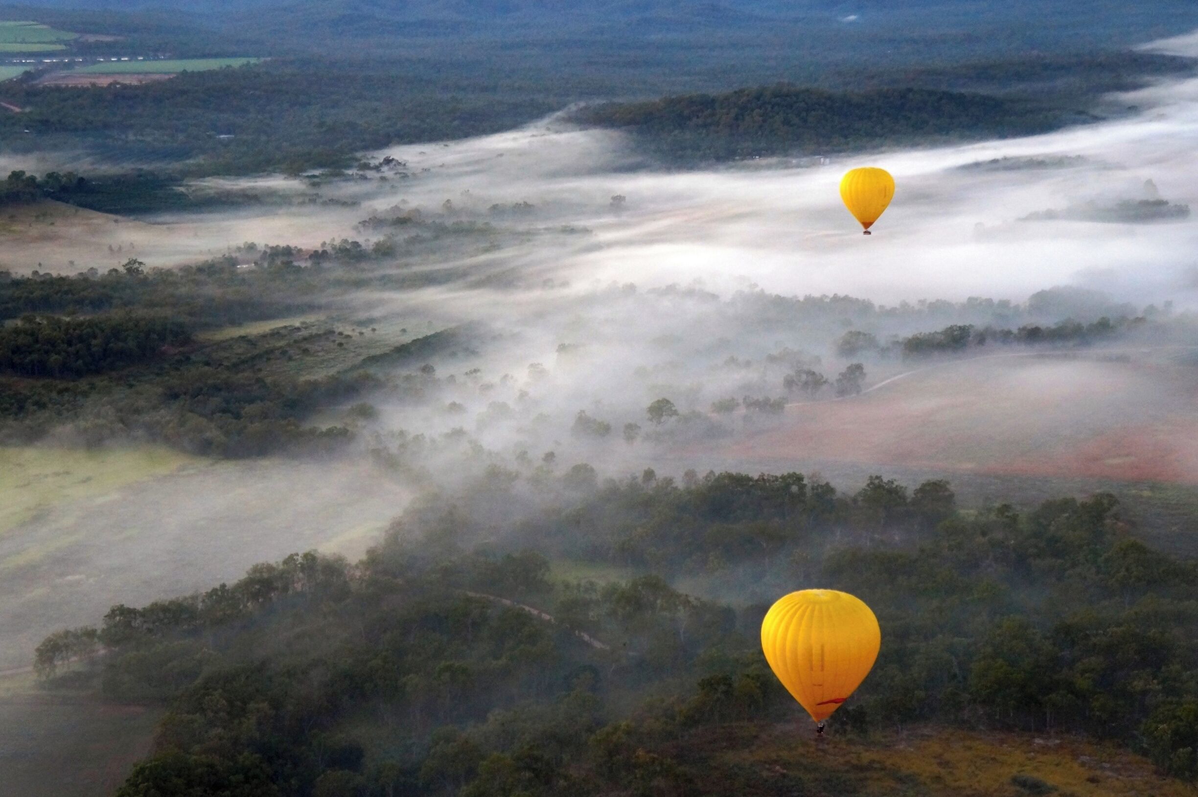 Two hot air balloons flying over a misty landscape with rolling hills and scattered trees.