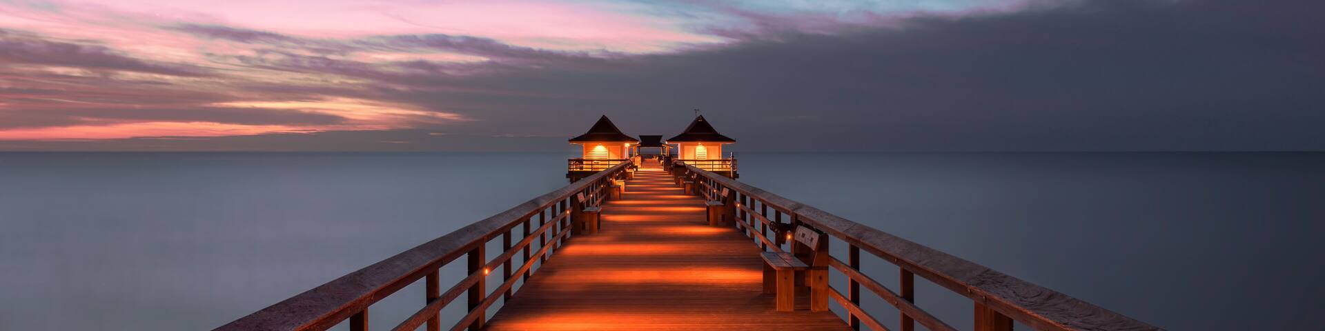 A wooden pier extending into calm waters at dusk, with two illuminated structures at the end.