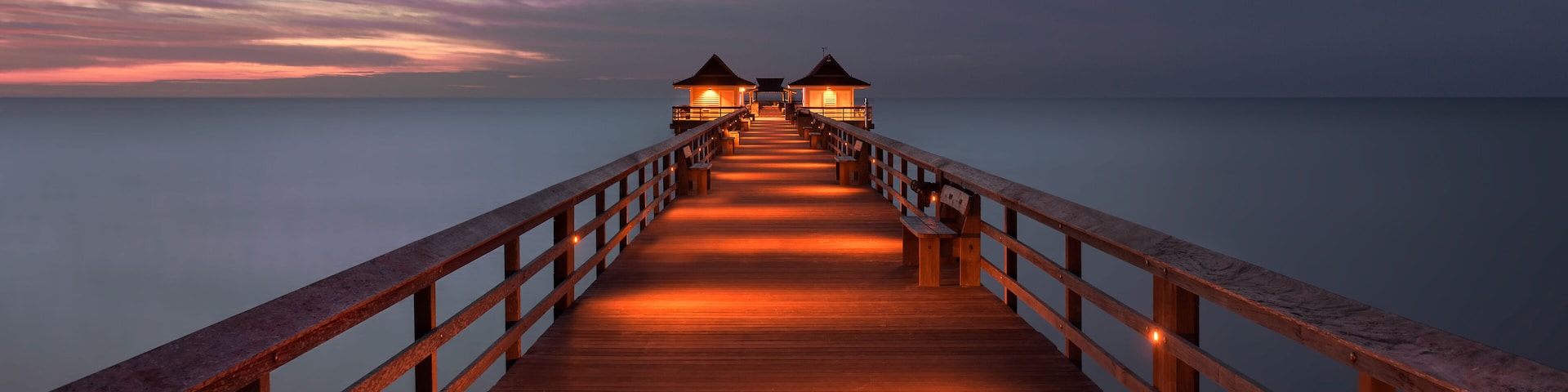 Une jetée en bois s’étendant sur des eaux calmes au crépuscule, avec deux structures illuminées à son extrémité.