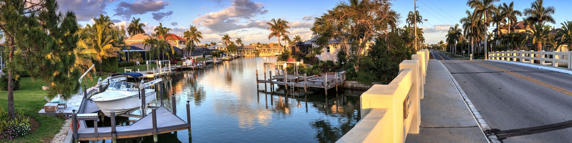 A marina with boats docked, a calm waterway, and palm trees lining the street.