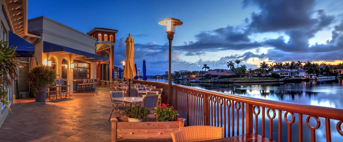 A waterfront patio with outdoor seating, a lamp post, and a view of the water and distant buildings.