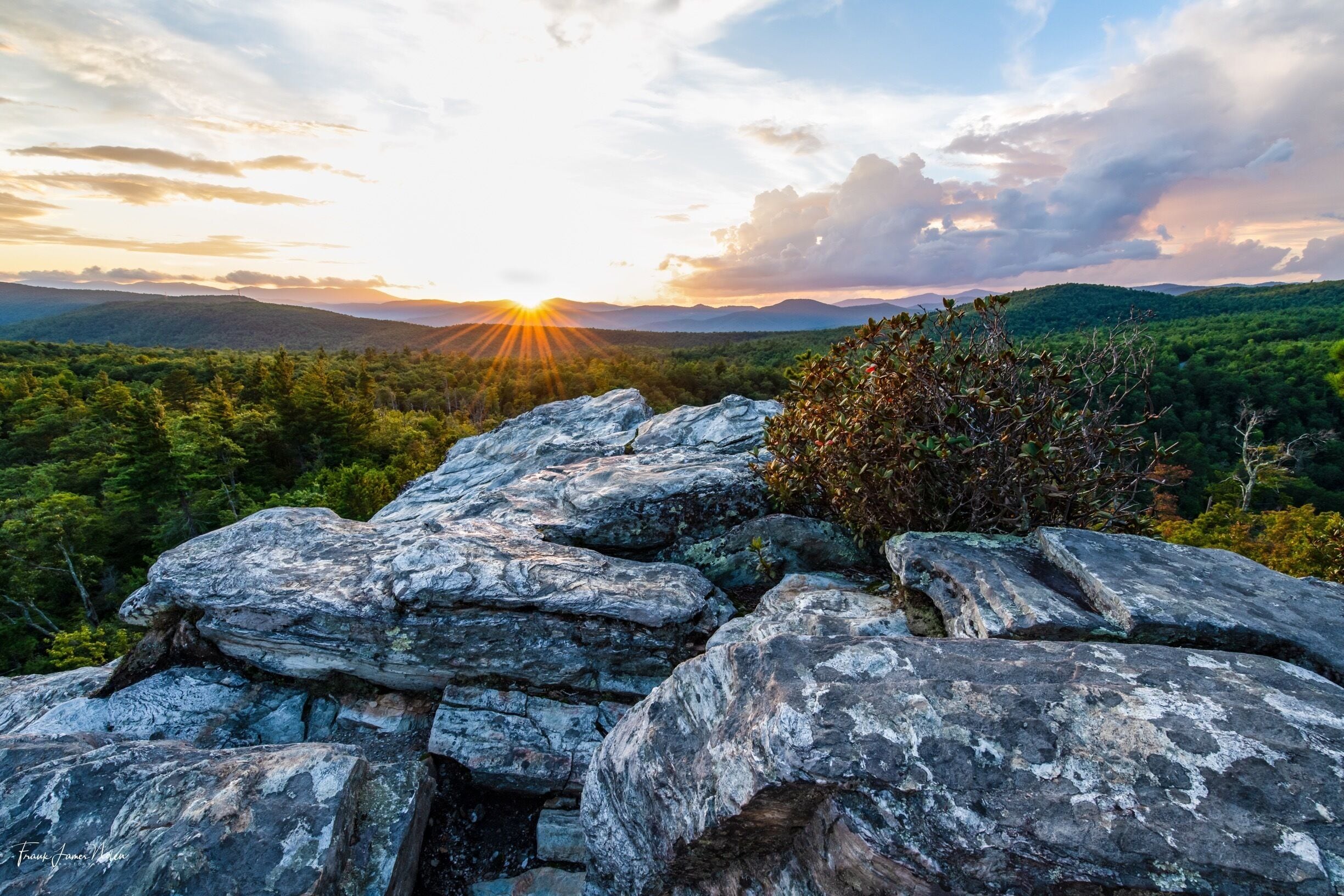 A rocky outcrop with a sunset over a forested mountain range.