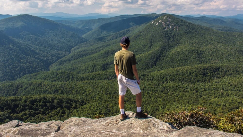 A person standing on a rock overlooking a mountainous landscape with dense forests.