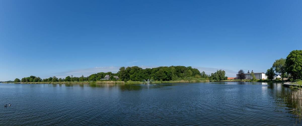 A calm lake with a fountain, surrounded by trees and buildings.
