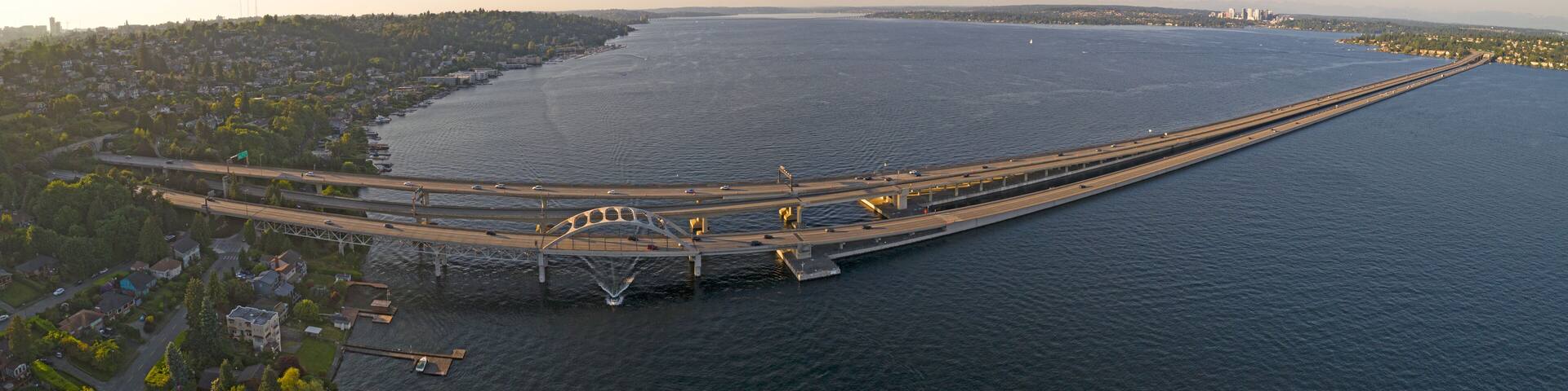 An aerial view of a large bridge spanning a wide river, with surrounding residential areas and distant cityscape.
