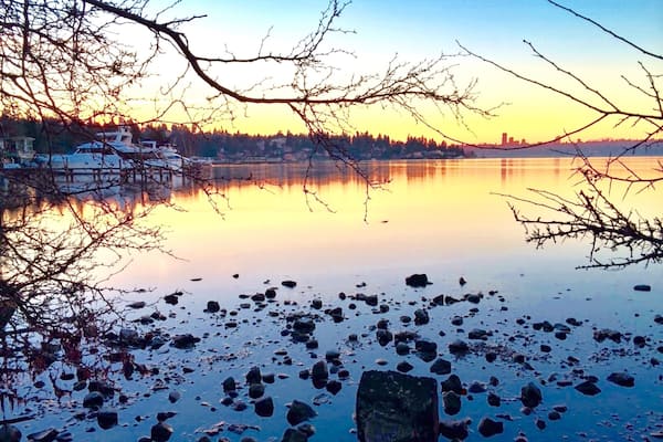 A calm lake with rocks in the foreground, a boat on the water, and a city skyline in the background during sunset.