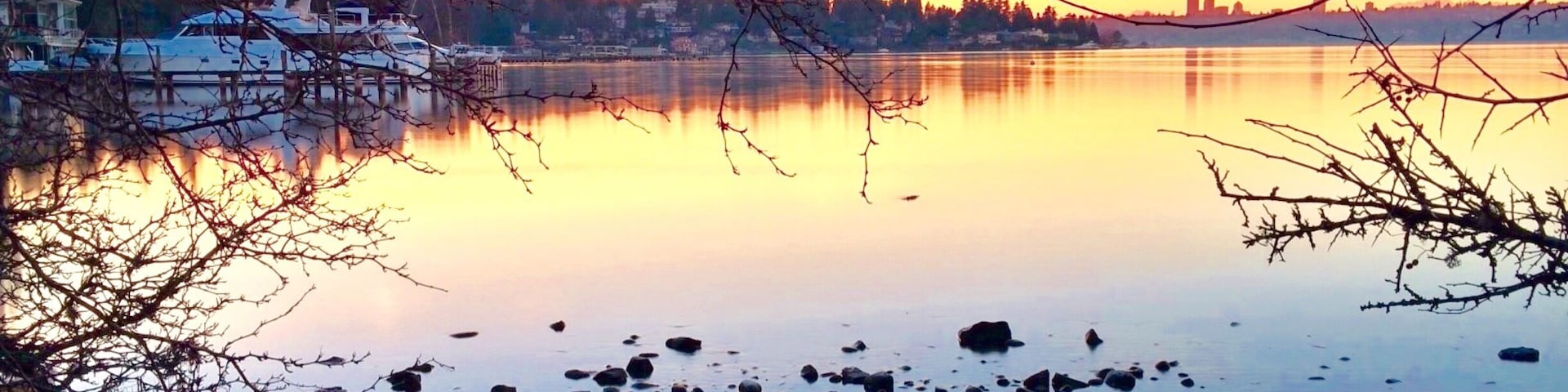 Um lago calmo com pedras em primeiro plano, um barco na água e um horizonte urbano ao fundo durante o pôr do sol.