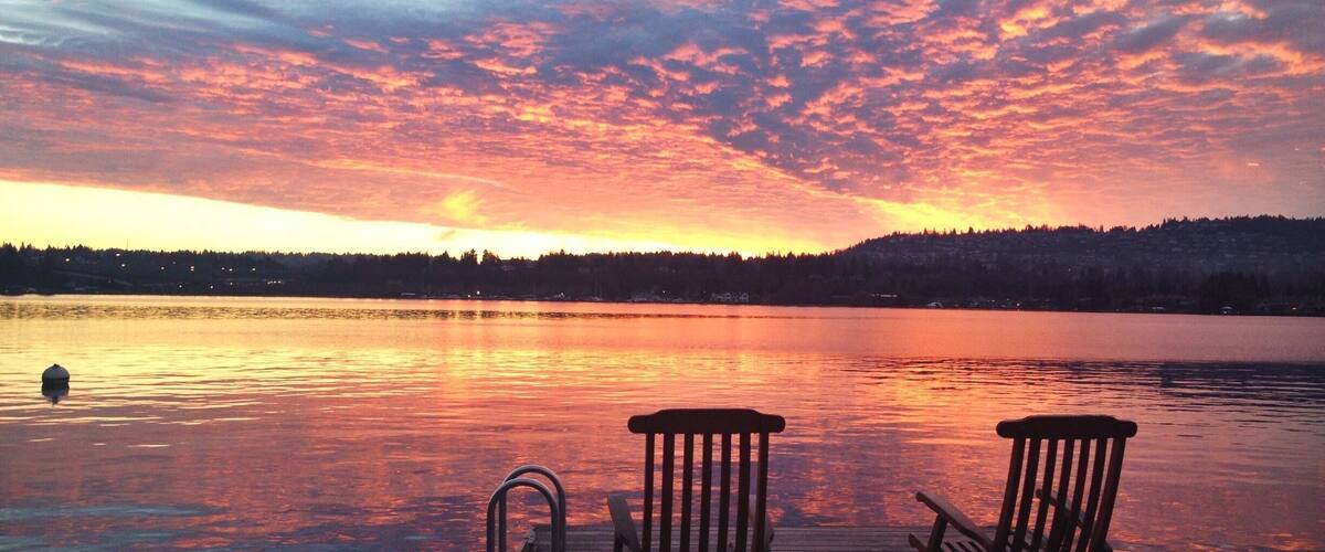 A sunset over a calm lake with two wooden chairs on a dock.