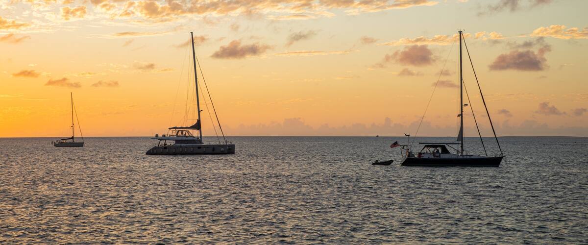 A sunset over calm waters with three sailboats.