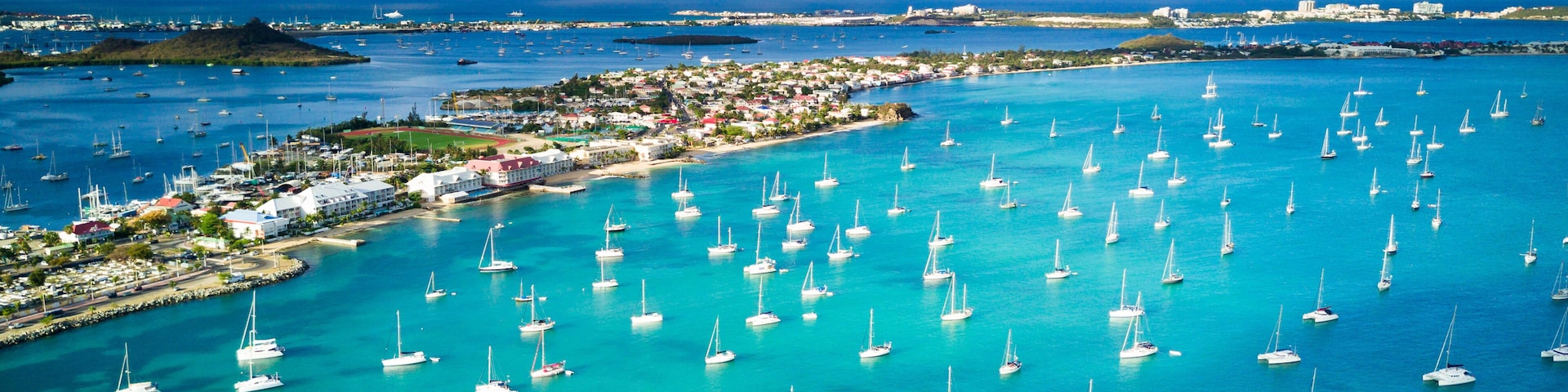 Un pueblo costero con numerosos veleros anclados en aguas turquesas, cielo despejado e islas en la distancia.