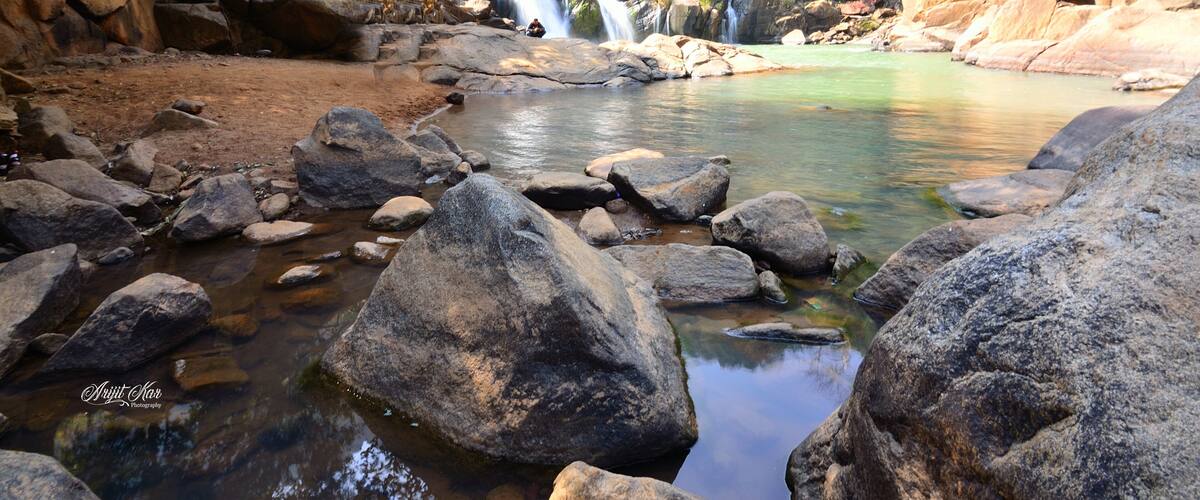 A rocky riverbed with clear water, large boulders, and a waterfall in a forested area.