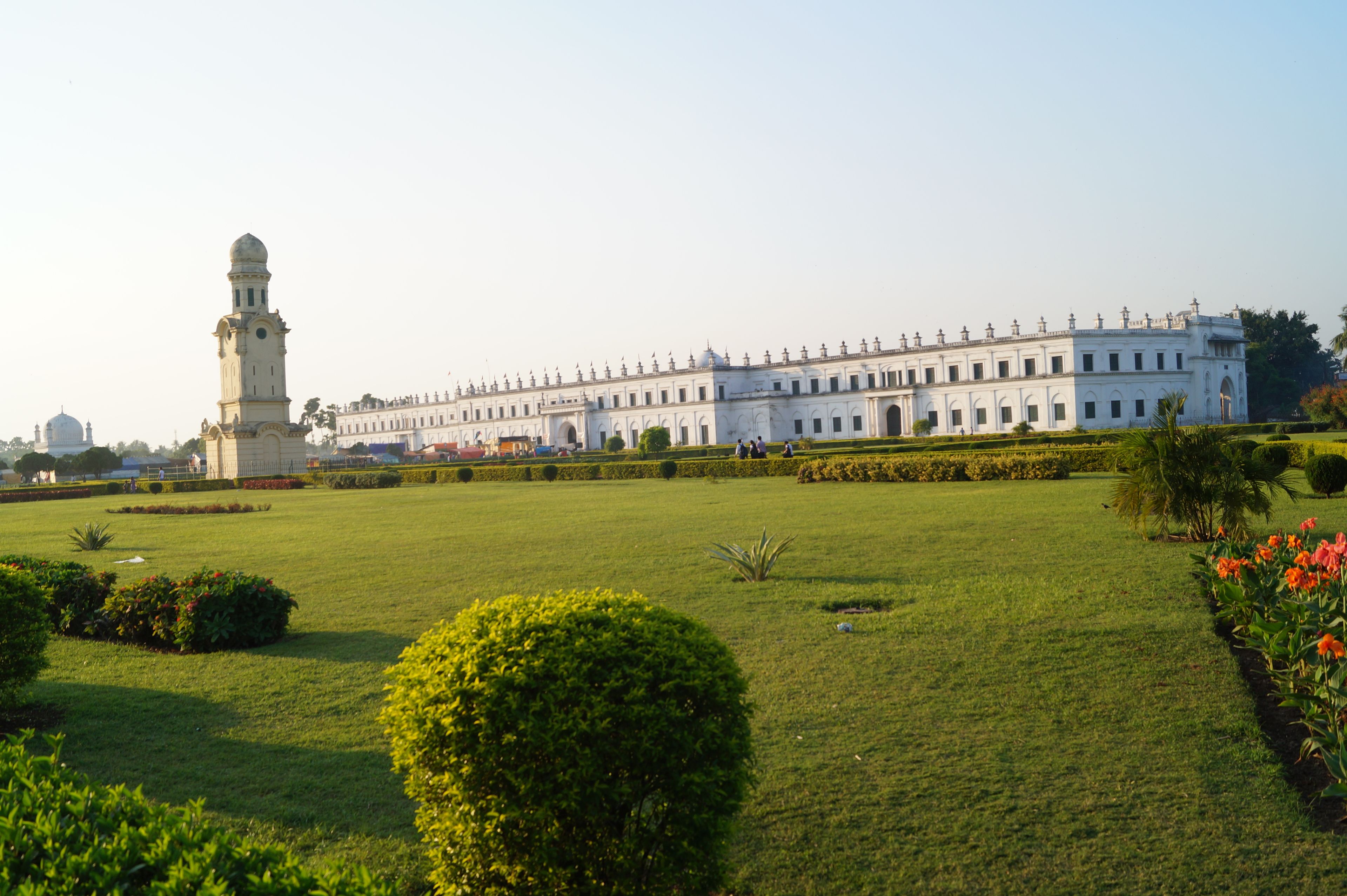 A large white building with a tower, surrounded by a well-maintained garden with green lawns and flowering plants.