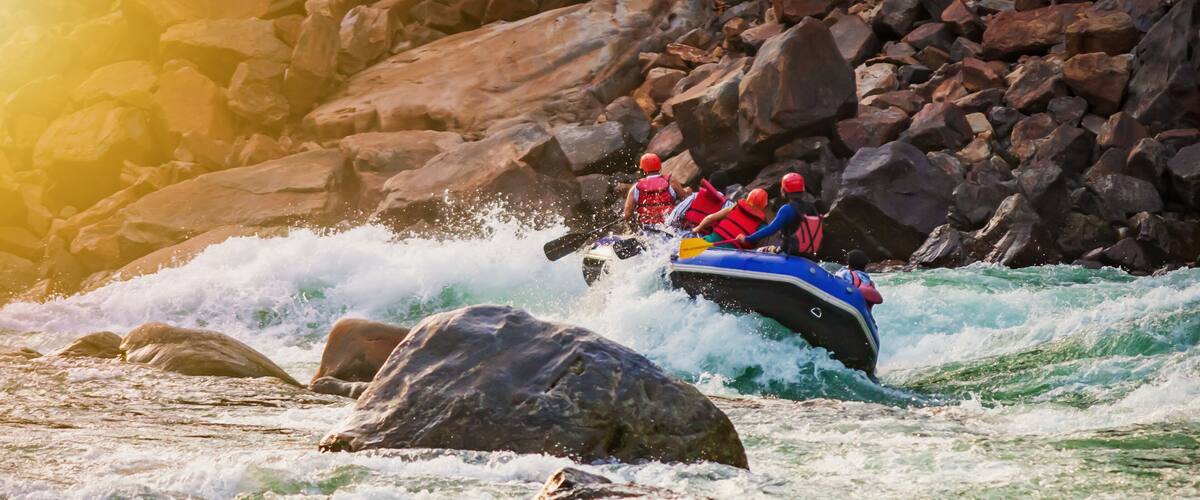 A group of people in a blue raft navigating through a river with large rocks and waves.