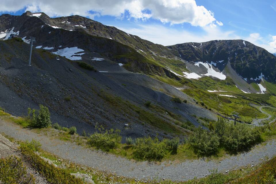 A mountain landscape with a winding path, patches of snow, and a distant lake.