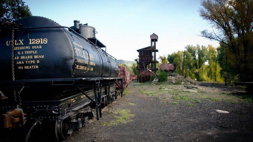 A black steam locomotive with the number 12918, labeled with technical specifications, traveling on tracks with a small building and trees in the background.