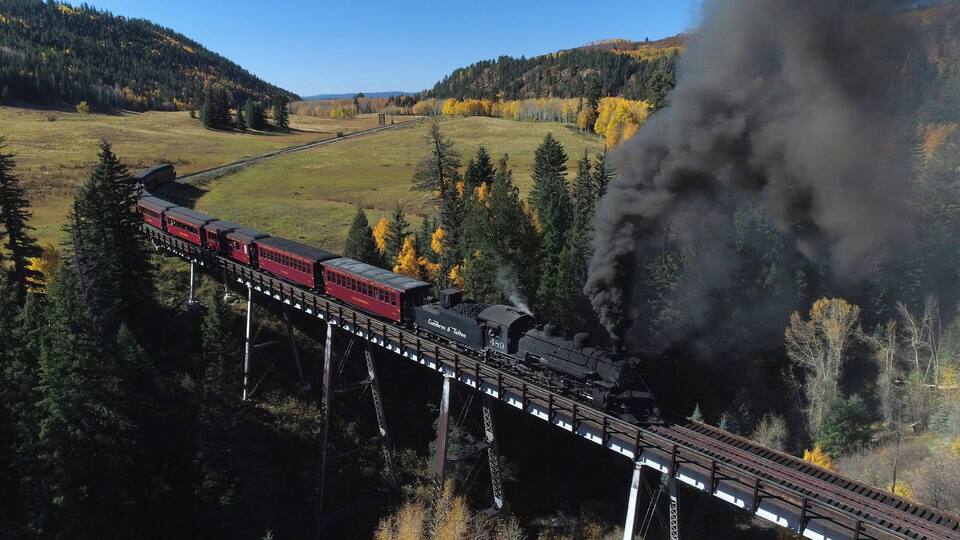 A steam locomotive pulling red passenger cars across a bridge over a forested valley.