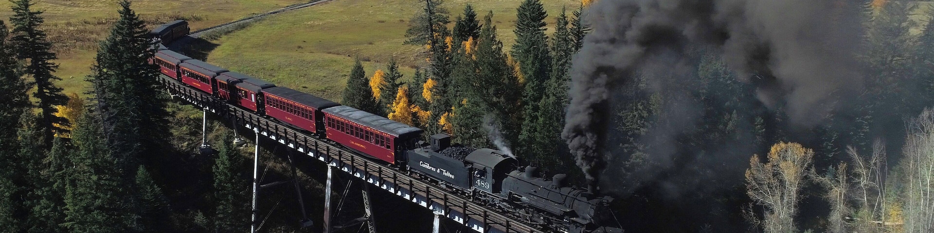 A steam locomotive pulling red passenger cars across a bridge over a forested valley.