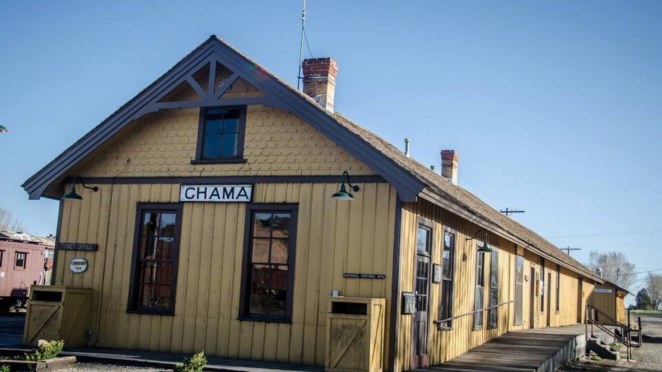 A wooden building labeled "CHAMA" with a ticket office sign, a small staircase, and a brick chimney.