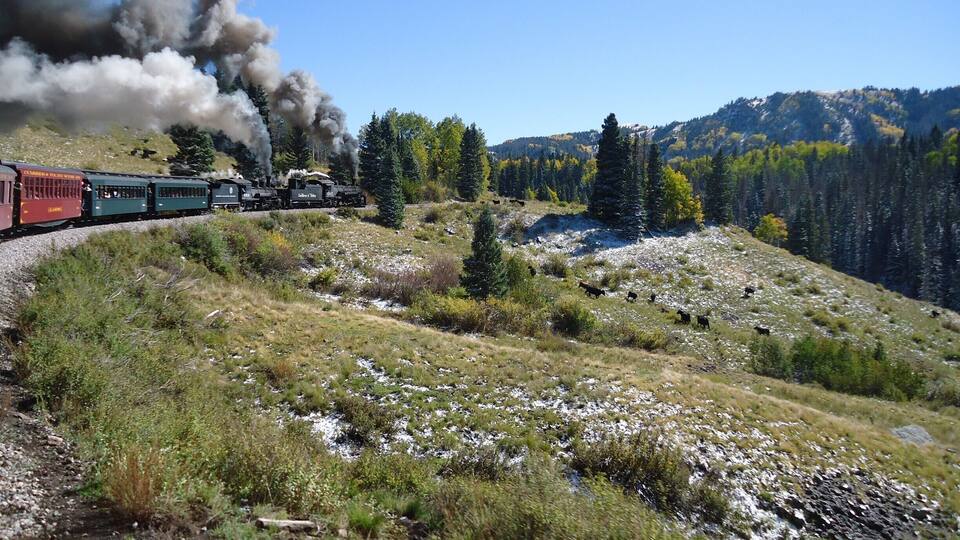 A train with black smoke on a mountain track surrounded by greenery and trees.