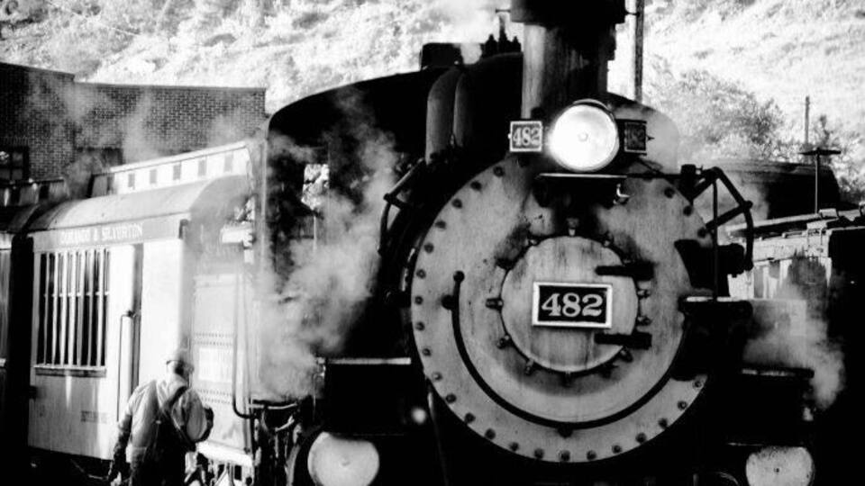 A steam locomotive numbered 482 at a railway station with a mountainous backdrop.