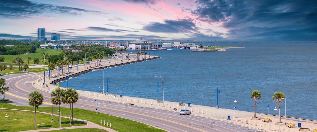 A coastal cityscape with a curved waterfront promenade, palm trees, and a distant bridge.