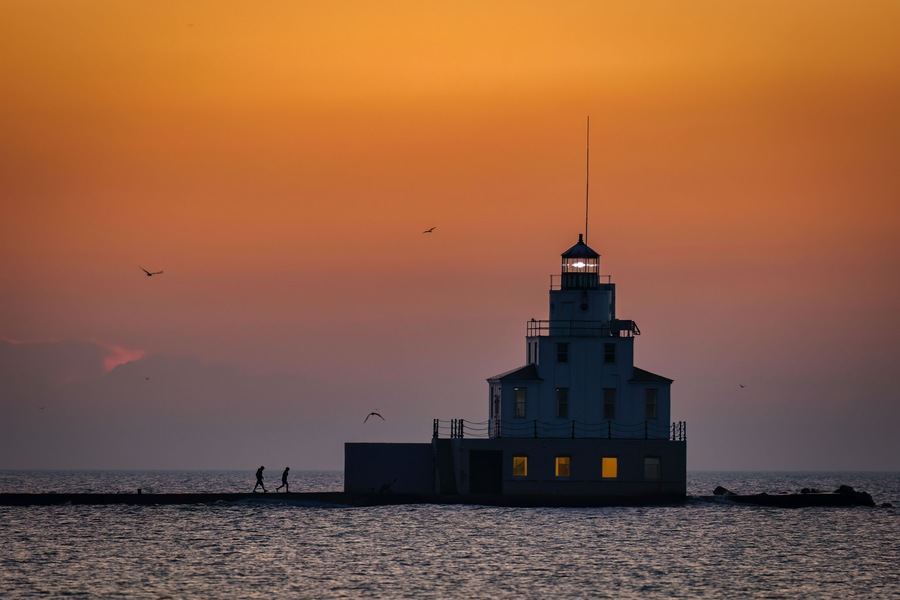 Un phare au coucher du soleil, avec les silhouettes de personnes et d’oiseaux.