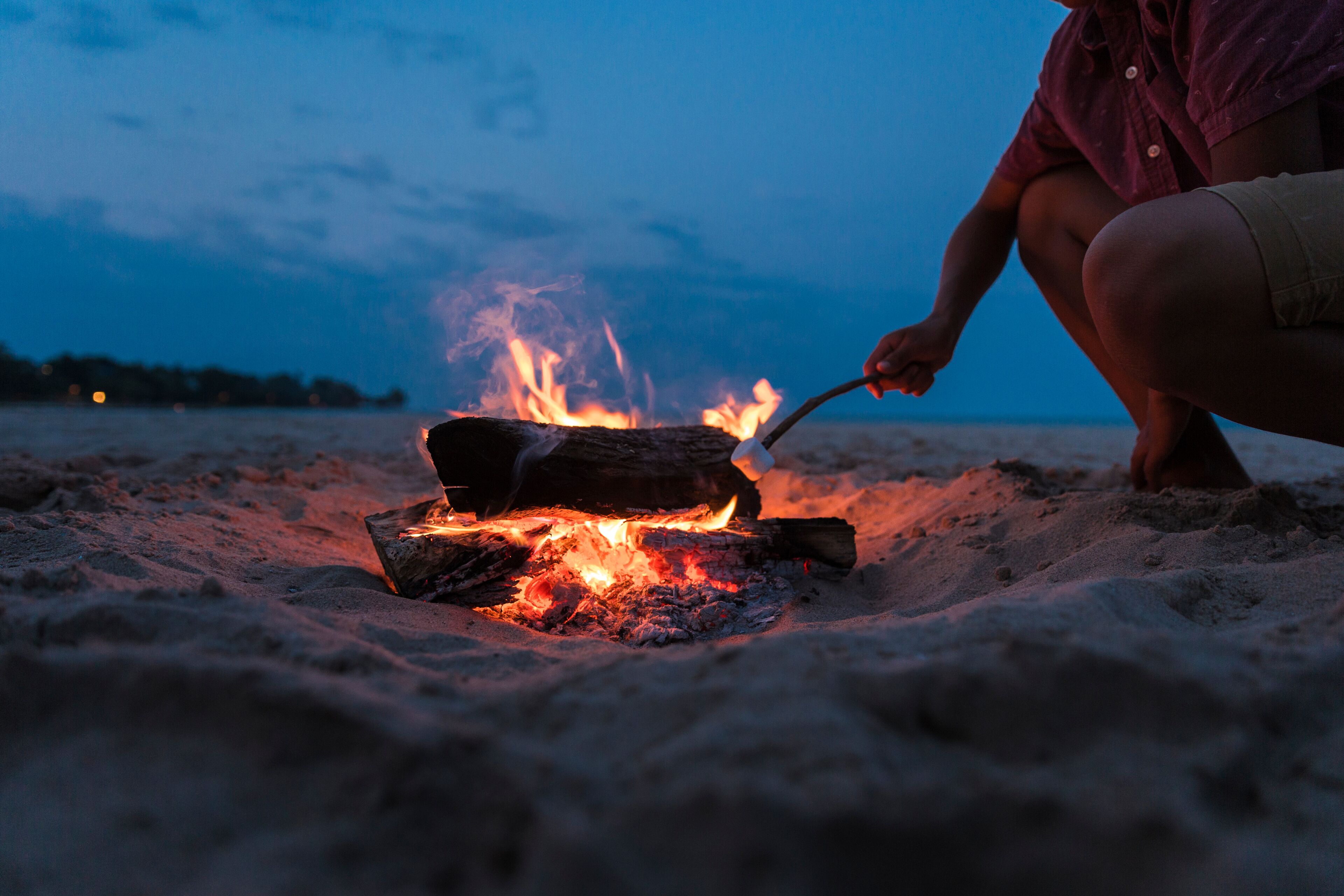 A person cooking over an open fire on a sandy beach at dusk.