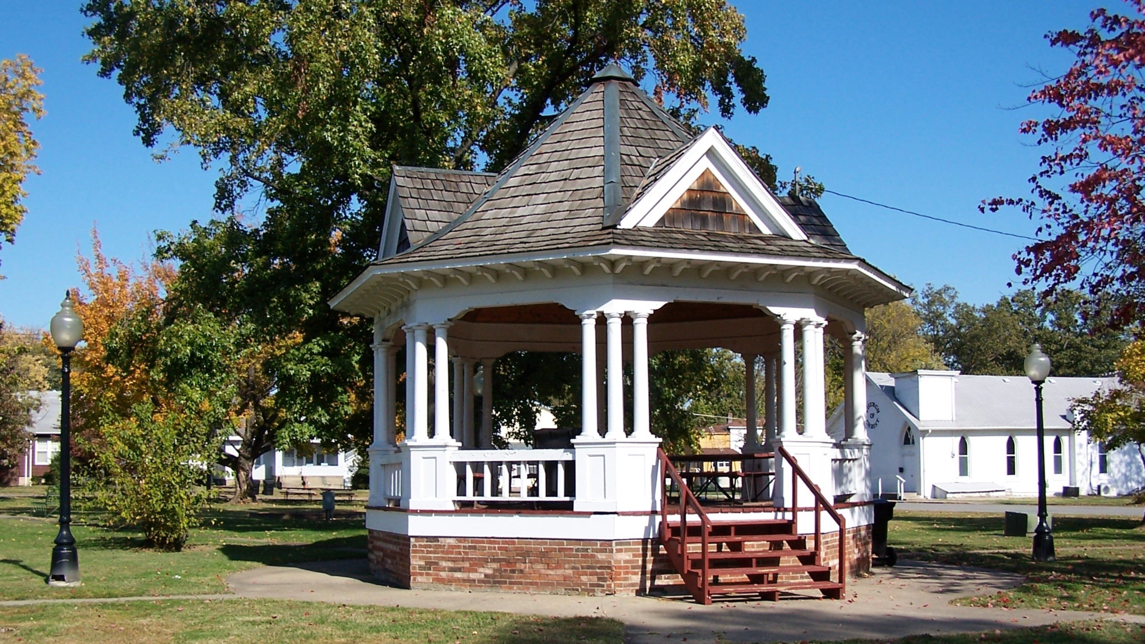 A gazebo with a peaked roof and white columns, surrounded by trees and a grassy area.