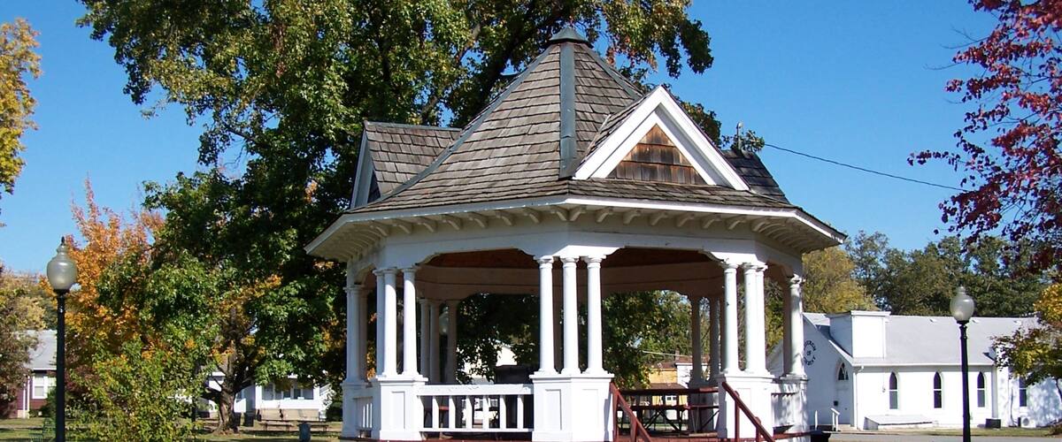 A gazebo with a peaked roof and white columns, surrounded by trees and a grassy area.