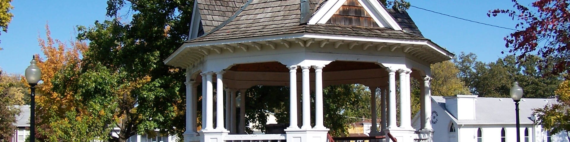 A gazebo with a peaked roof and white columns, surrounded by trees and a grassy area.