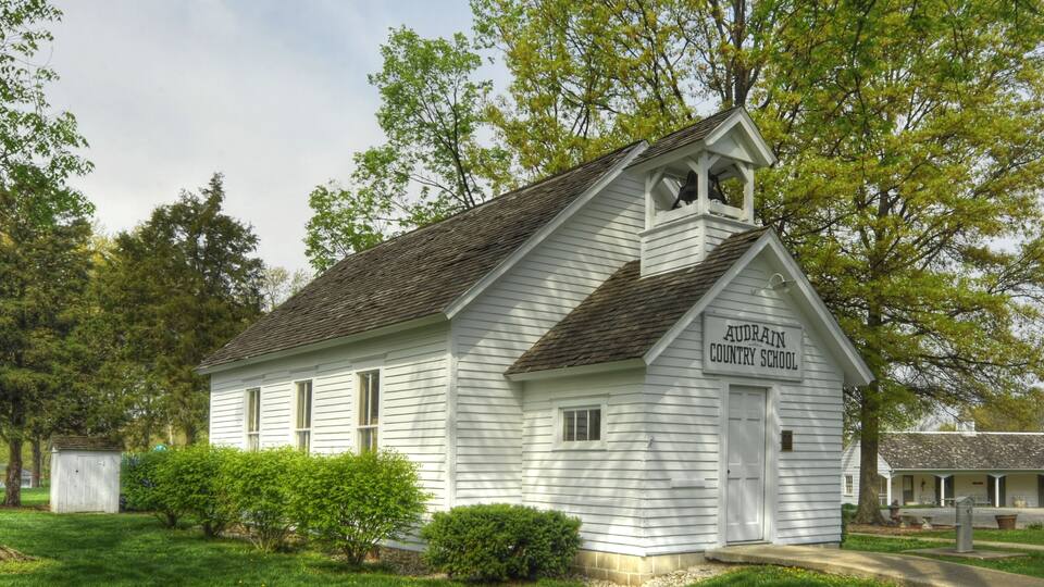 A white wooden schoolhouse with a bell tower, surrounded by lush green trees and a well-maintained lawn.