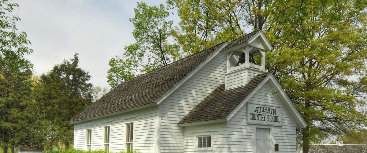 A white wooden schoolhouse with a bell tower, surrounded by lush green trees and a well-maintained lawn.