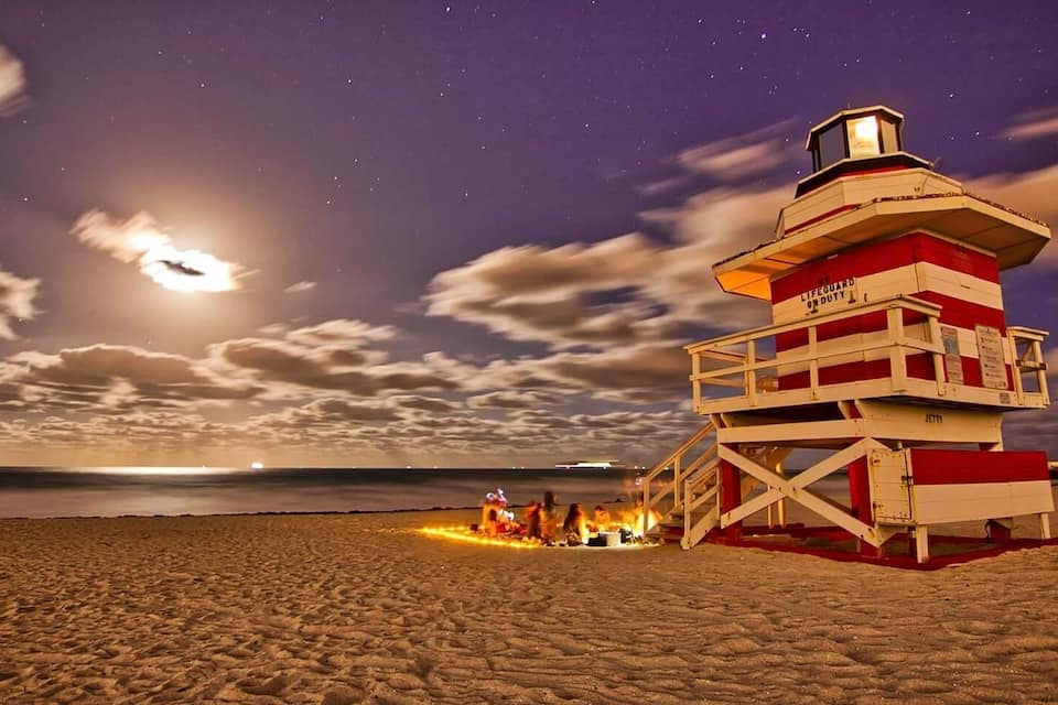 A beach with a red and white lifeguard tower and a bonfire.