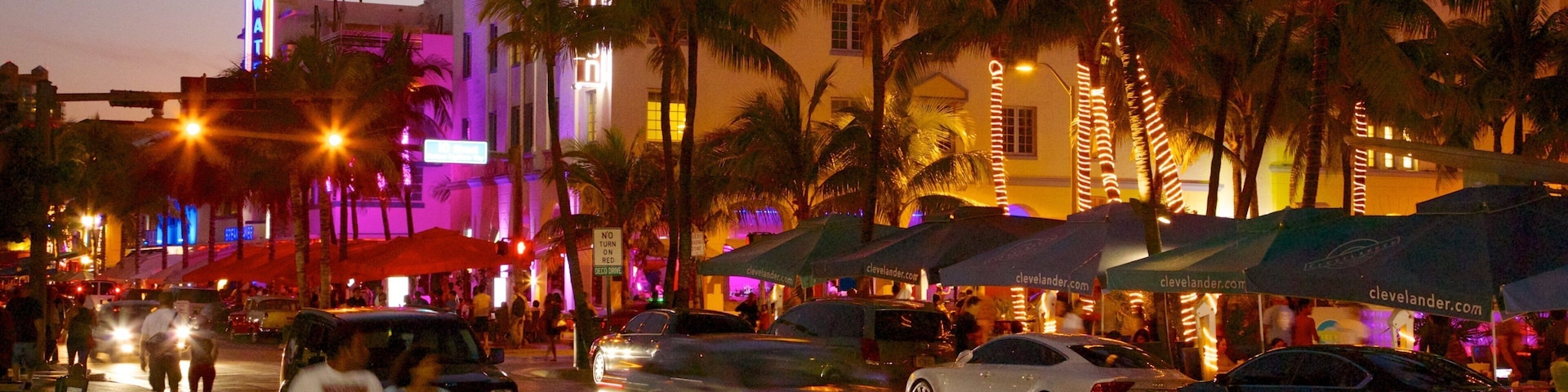 Uma rua movimentada ao entardecer, com palmeiras, carros e edifícios iluminados.